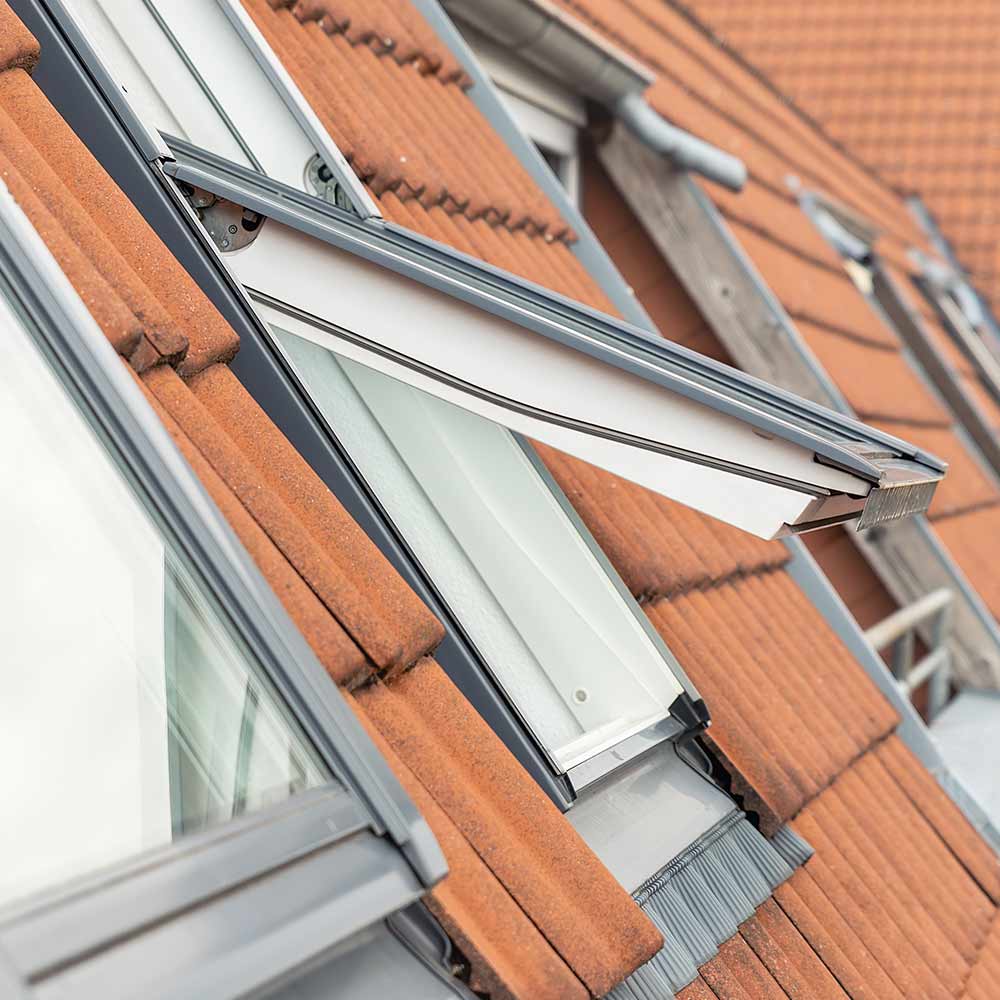 Fenêtre de toit Velux ouverte en partie haute, s'intégrant dans un toit en tuiles rouges, avec châssis gris moderne.