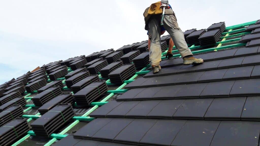 Pose de Tuiles Noires : Chantier de Couverture en Cours Couvreur au travail sur un toit en pente, posant des tuiles plates noires sur des liteaux verts, avec des palettes de tuiles prêtes à être installées.