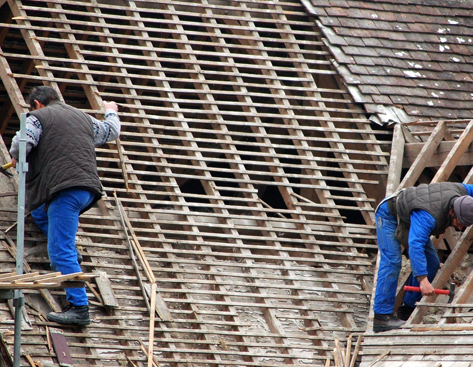 Deux couvreurs vêtus de bleu et de gilets matelassés rénovent une vieille toiture, retirant les vieilles tuiles pour exposer le lattis en bois.