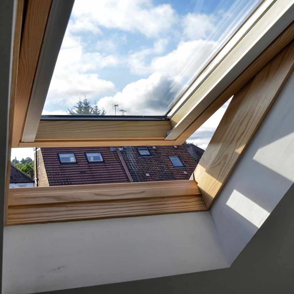 Velux en bois clair ouvert montrant un ciel bleu nuageux et des toits de tuiles rouges avec d'autres fenêtres de toit